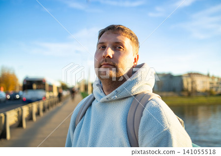 man looking up in urban riverside setting with thoughtful expression and backpack straps visible on concrete road near metal railing, exploring city life and travel lifestyle. man looking up in urban riverside setting with thoughtful expression and backpack straps visible on concrete road near metal railing, exploring city life and travel lifestyle. 141055518
