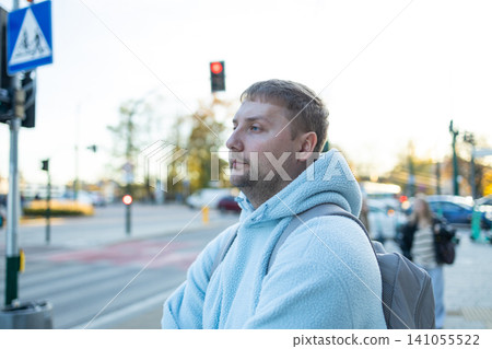 man waiting at a crosswalk in urban street scene with traffic lights and pedestrians, side view, neutral mood, blue jacket, concrete pavement, gray housing, everyday life, public transport. man waiting at a crosswalk in urban street scene with traffic lights and pedestrians, side view, neutral mood, blue jacket, concrete pavement, gray housing, everyday life, public transport. 141055522