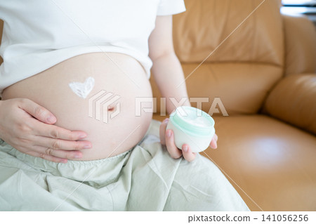 Pregnant woman applying heart shaped stretch mark cream to her belly while sitting on the sofa at home. Preganacy body care routine concept. 141056256