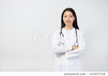 A young Japanese female doctor in a white coat stands with her arms crossed in front of a white background, smiling and looking directly at the camera. A young Japanese female doctor in a white coat stands with her arms crossed in front of a white background, smiling and looking directly at the camera. 141057148