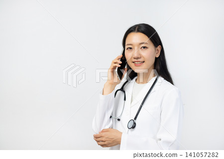 A young Japanese female doctor in a white coat smiles at the camera, holding a smartphone to her ear in front of a white background. A young Japanese female doctor in a white coat smiles at the camera, holding a smartphone to her ear in front of a white background. 141057282