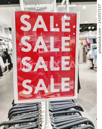 Red Sale Sign In Clothing Store Over Racks Advertising Discount Promotion And Seasonal Retail Savings. Red Sale Sign In Clothing Store Over Racks Advertising Discount Promotion And Seasonal Retail Savings. 141057337