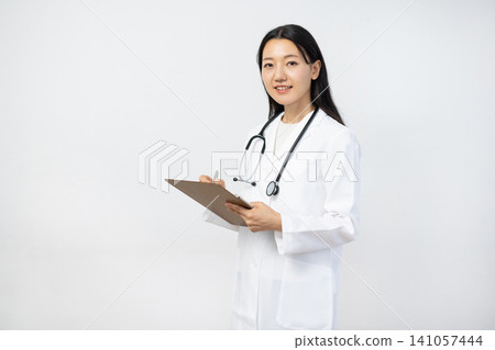 A young Japanese female doctor in a white coat smiles as she takes notes in a binder in front of a white background. A young Japanese female doctor in a white coat smiles as she takes notes in a binder in front of a white background. 141057444
