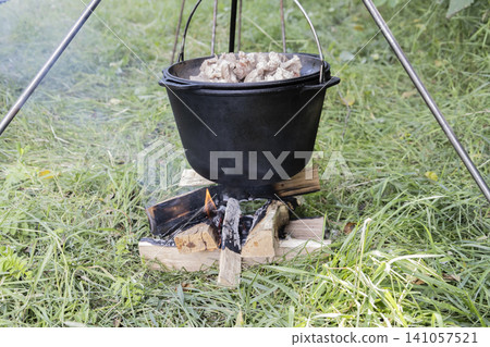 Cooking delicious meat in a large cast-iron cauldron over an open campfire in nature. A rustic outdoor meal preparation scene, showcasing traditional cooking methods during a hike or camping Cooking delicious meat in a large cast-iron cauldron over an open campfire in nature. A rustic outdoor meal preparation scene, showcasing traditional cooking methods during a hike or camping 141057521