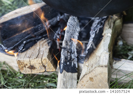 Close-up view of a small campfire made of stacked wood logs and charcoal on green grass, actively burning with visible flames and rising smoke, heating the bottom of a black pot for cooking. Close-up view of a small campfire made of stacked wood logs and charcoal on green grass, actively burning with visible flames and rising smoke, heating the bottom of a black pot for cooking. 141057524