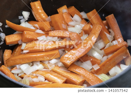Sliced fresh carrots and white onions roasting in a heavy cast-iron cauldron over an outdoor campfire. A rustic, healthy meal being prepared during a camping trip, showcasing natural cooking Sliced fresh carrots and white onions roasting in a heavy cast-iron cauldron over an outdoor campfire. A rustic, healthy meal being prepared during a camping trip, showcasing natural cooking 141057542