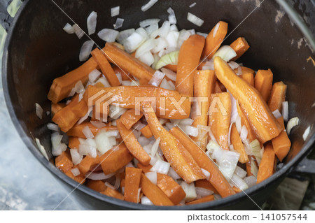 Sliced fresh carrots and white onions roasting in a heavy cast-iron cauldron over an outdoor campfire. A rustic, healthy meal being prepared during a camping trip, showcasing natural cooking Sliced fresh carrots and white onions roasting in a heavy cast-iron cauldron over an outdoor campfire. A rustic, healthy meal being prepared during a camping trip, showcasing natural cooking 141057544