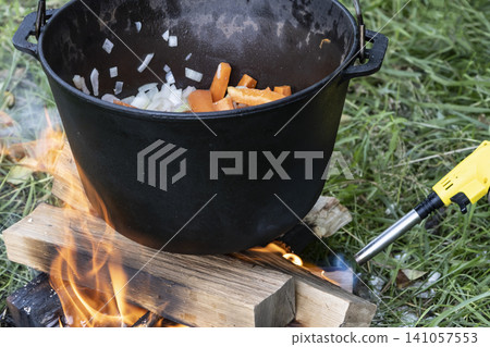 Black cast iron pot with carrots and onions, hanging over a small campfire built on the green grass. Perfect for rustic outdoor cooking and camping. Black cast iron pot with carrots and onions, hanging over a small campfire built on the green grass. Perfect for rustic outdoor cooking and camping. 141057553