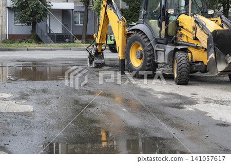 Excavator hydraulic hammer breaking asphalt on a street with puddles and passing traffic. Heavy machinery repairing road surface in urban area with trees and residential building in background. Excavator hydraulic hammer breaking asphalt on a street with puddles and passing traffic. Heavy machinery repairing road surface in urban area with trees and residential building in background. 141057617