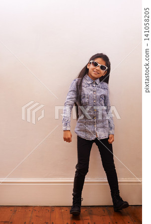 Girl, child and fashion or sunglasses for style and looking cool on a wall background at home. Happy portrait of a kid or child with trendy clothes, casual and accessory or shades for fun indoors 141058205