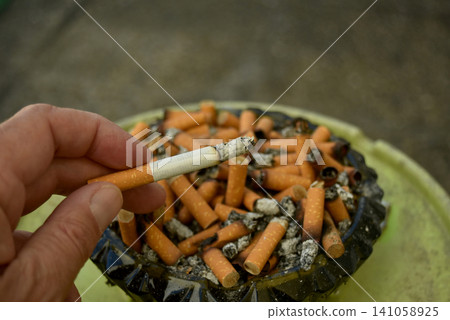 Closeup of a hand holding a burning cigarette above an ashtray filled with cigarette butts and ashes. Concept of smoking, nicotine addiction, tobacco consumption and public health. Closeup of a hand holding a burning cigarette above an ashtray filled with cigarette butts and ashes. Concept of smoking, nicotine addiction, tobacco consumption and public health. 141058925