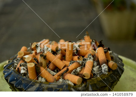 Closeup of an ashtray filled with cigarette butts and ashes. Concept of tobacco consumption, nicotine addiction, smoking waste and public health awareness. Closeup of an ashtray filled with cigarette butts and ashes. Concept of tobacco consumption, nicotine addiction, smoking waste and public health awareness. 141058979