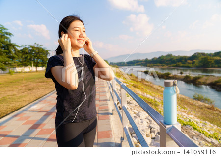 Woman wearing earphones preparing for outdoor exercise near river. Fitness and healthy lifestyle concept with workout, wellness and active living. Woman wearing earphones preparing for outdoor exercise near river. Fitness and healthy lifestyle concept with workout, wellness and active living. 141059116