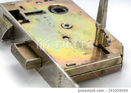 Detailed closeup of vintage brass mortise door lock mechanism with antique key inserted into keyhole showing weathered metal surface, screws, latch bolt and internal locking components on white Detailed closeup of vintage brass mortise door lock mechanism with antique key inserted into keyhole showing weathered metal surface, screws, latch bolt and internal locking components on white 141059584