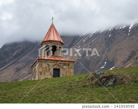 Ancient Stone Bell Tower of Gergeti Trinity Church Against Cloudy Caucasus Mountains in Kazbegi, Georgia. Ancient Stone Bell Tower of Gergeti Trinity Church Against Cloudy Caucasus Mountains in Kazbegi, Georgia. 141059595