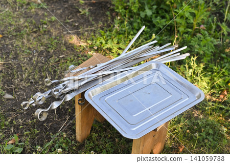 Metal skewers and tray for barbecue placed on small wooden stand outdoors, closeup of cooking tools ready for grilling, picnic and outdoor culinary concept Metal skewers and tray for barbecue placed on small wooden stand outdoors, closeup of cooking tools ready for grilling, picnic and outdoor culinary concept 141059788