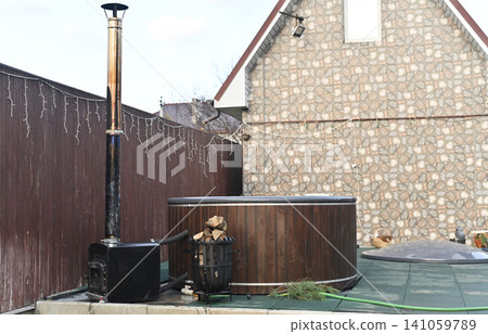 Outdoor hot tub with wood exterior and metal stove beside fence, installed on terrace near brick building. Hot tub heated with firewood creating cozy spa atmosphere, backyard relaxation concept, 141059789