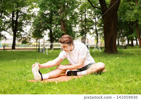 man is stretching on a mat on green grass in the park. This outdoor fitness exercise and daily workout routine is excellent for his overall health. man is stretching on a mat on green grass in the park. This outdoor fitness exercise and daily workout routine is excellent for his overall health. 141059930