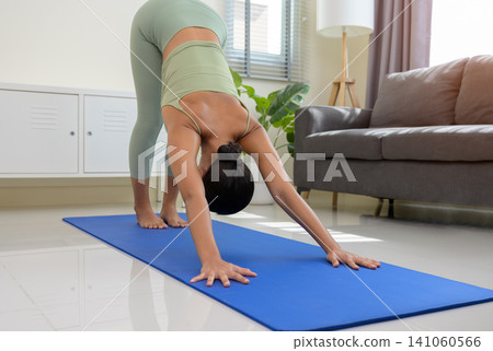 Young asian woman practicing yoga in downward facing dog pose on blue mat in cozy living room at home Young asian woman practicing yoga in downward facing dog pose on blue mat in cozy living room at home 141060566