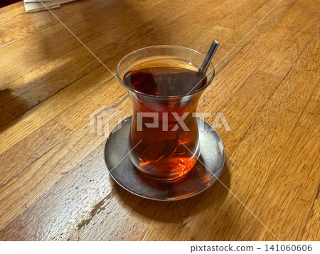 Traditional Turkish tea served in tulip shaped glass on metal saucer with spoon over wooden table. Turkish culture, hospitality, beverage, cafe, lifestyle, travel, food culture, refreshment 141060606