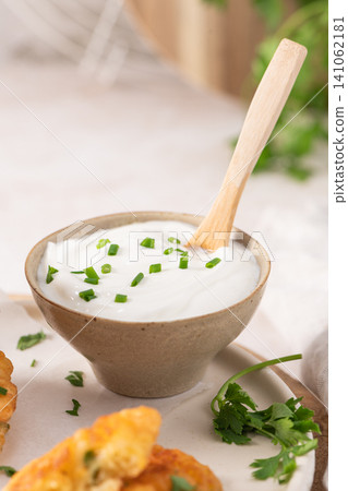 A small ceramic bowl of creamy yogurt or savory sour cream topped with fresh chopped green chives and a wooden spoon, served alongside crispy fritters A small ceramic bowl of creamy yogurt or savory sour cream topped with fresh chopped green chives and a wooden spoon, served alongside crispy fritters 141062181