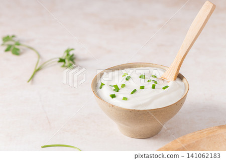A ceramic bowl of creamy white yogurt or savory sour cream topped with fresh chopped green chives, served with a wooden spoon on a light surface 141062183
