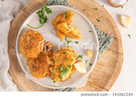 Crispy golden cod fritters served on a plate with fresh parsley, lemon wedges, and sea salt, arranged on a rustic wooden board with a soft fabric Crispy golden cod fritters served on a plate with fresh parsley, lemon wedges, and sea salt, arranged on a rustic wooden board with a soft fabric 141062184