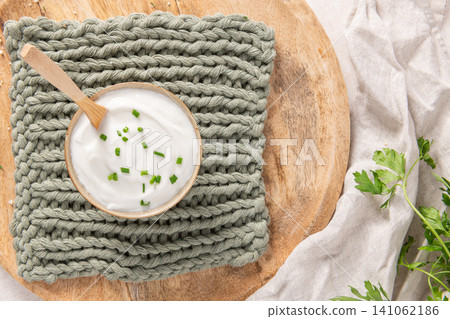 A bowl of creamy white yogurt or sour cream topped with fresh chives sits on a rustic wooden board and a soft green knit cloth with a casual fabric drape 141062186
