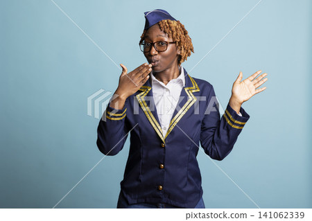African american air hostess blows kiss while smiling in studio with blue background. Female cabin crew member covers mouth with hand, acting surprised over impressive flight deals. African american air hostess blows kiss while smiling in studio with blue background. Female cabin crew member covers mouth with hand, acting surprised over impressive flight deals. 141062339