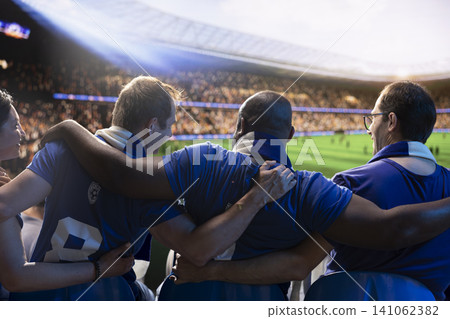 Rear view of soccer supporters sitting shoulder to shoulder with arms around each other in packed stadium. Crowd doing choreography in sports arena stands, showing team support Rear view of soccer supporters sitting shoulder to shoulder with arms around each other in packed stadium. Crowd doing choreography in sports arena stands, showing team support 141062382