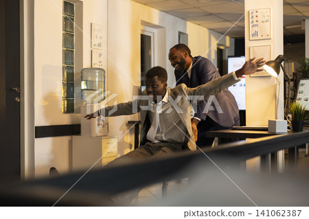 Businessman giving colleague joyful ride on chair in startup office after hours. Silly excited people enjoying playful office games during overtime, positive team connection and stress relief. Businessman giving colleague joyful ride on chair in startup office after hours. Silly excited people enjoying playful office games during overtime, positive team connection and stress relief. 141062387