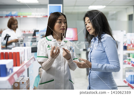 Asian pharmacist assists curious female customer browsing supplements in local retail pharmacy. Professional consultation focuses on product benefits, wellbeing and accurate health information. Asian pharmacist assists curious female customer browsing supplements in local retail pharmacy. Professional consultation focuses on product benefits, wellbeing and accurate health information. 141062389