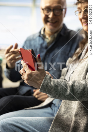 Close up of modern active retirees using mobile phone to check travel itinerary and sightseeing list, sitting in airport lounge. Old couple representing unconventional lifestyle in later life. Close up of modern active retirees using mobile phone to check travel itinerary and sightseeing list, sitting in airport lounge. Old couple representing unconventional lifestyle in later life. 141062392
