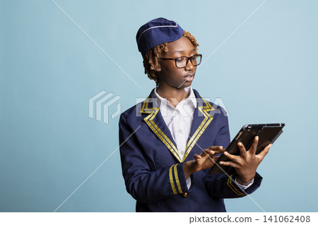 Focused black woman in airline uniform standing confidently and holding tablet. Female flight attendant reviewing passenger information and preparing for upcoming travel operations digitally. 141062408