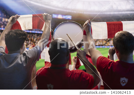 Soccer fans in stadium tribunes playing drum during intense soccer match, creating passionate atmosphere. Football supporters raising red team kit scarves to support players on pitch Soccer fans in stadium tribunes playing drum during intense soccer match, creating passionate atmosphere. Football supporters raising red team kit scarves to support players on pitch 141062413
