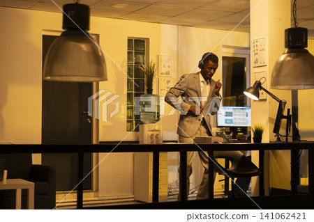 Black businessman, silly dancing moves, office workspace, late at night. After hours office contrasts with carefree leisure moment, employee enjoying groovy music with positive energy. Black businessman, silly dancing moves, office workspace, late at night. After hours office contrasts with carefree leisure moment, employee enjoying groovy music with positive energy. 141062421