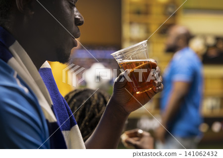 Close up of football supporter drinking beer from plastic cup while live match plays on TV in living room. Side view of man enjoying alcohol beverage during relaxed home watch party Close up of football supporter drinking beer from plastic cup while live match plays on TV in living room. Side view of man enjoying alcohol beverage during relaxed home watch party 141062432