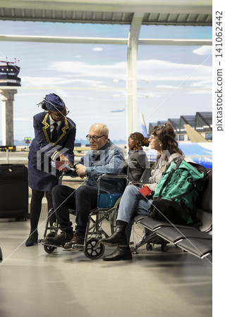 Smiling cabin crew member assisting elderly passenger with mobility aid, checking boarding pass and giving directions at airport terminal. Senior couple represents inclusive tourism. 141062442