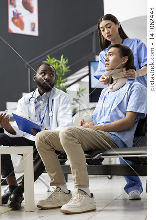 Young adult man sits with black male physician in hospital lobby, listening attentively. Doctor explains medical procedures as nurse adjusts neck brace on patient after diagnostic tests in clinic 141062443