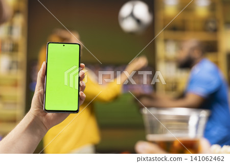 Green screen phone fills foreground while football match plays on blurred television set. Football supporters use mockup phone at home during live game broadcast coverage Green screen phone fills foreground while football match plays on blurred television set. Football supporters use mockup phone at home during live game broadcast coverage 141062462