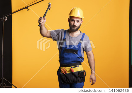 Male renovator in yellow hard hat lifts hammer with firm grip, showing readiness for repair or building task. Confident handyman stands in blue overalls and tool belt against isolated background. Male renovator in yellow hard hat lifts hammer with firm grip, showing readiness for repair or building task. Confident handyman stands in blue overalls and tool belt against isolated background. 141062486