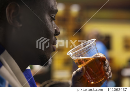 Close up of football fan drinking beer while watching soccer match in living room. Man enjoying refreshments during evening live sports game event Close up of football fan drinking beer while watching soccer match in living room. Man enjoying refreshments during evening live sports game event 141062487