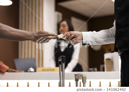 Close up of traveler giving money banknotes to bellboy near front desk, help with heavy suitcases. Hospitality industry scene emphasizes payment and value of services. Gratitude for hotel staff. Close up of traveler giving money banknotes to bellboy near front desk, help with heavy suitcases. Hospitality industry scene emphasizes payment and value of services. Gratitude for hotel staff. 141062492