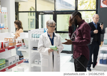 Drugstore attendant advises black man on suitable medication for headache relief at local pharmacy. Male customer consults about pills for headache and allergy symptoms during healthcare interaction. 141062505
