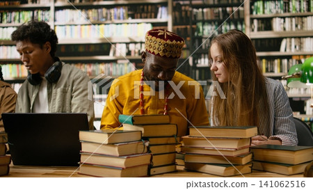 Students happily discussing study materials and reading from textbooks at shared library table, collaborative work during group study surrounded by green lamps and bookshelves. Students happily discussing study materials and reading from textbooks at shared library table, collaborative work during group study surrounded by green lamps and bookshelves. 141062516