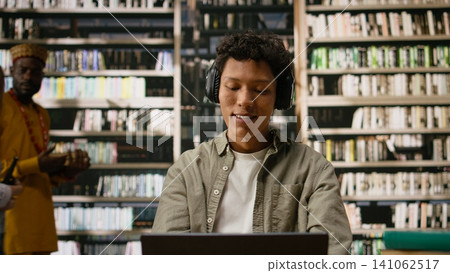 Student with headphones reading open book and studying with focus music, multitasking at desk with laptop and study materials during study session with peers. Quiet library space. 141062517