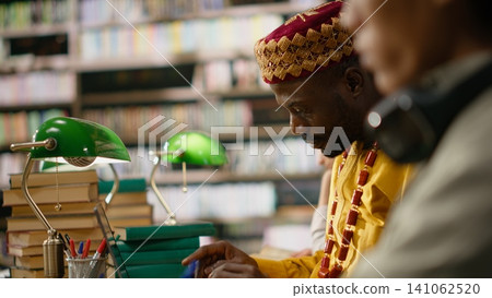 African mature student in yellow traditional clothing smiles while working on laptop at desk in study area with stacks of books, monitors and classmates nearby. Digital learning. African mature student in yellow traditional clothing smiles while working on laptop at desk in study area with stacks of books, monitors and classmates nearby. Digital learning. 141062520