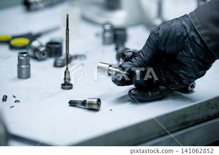 Mechanic hand in glove holding new fuel injector nozzle replacement part near old dirty disassembled components in workshop Mechanic hand in glove holding new fuel injector nozzle replacement part near old dirty disassembled components in workshop 141062852