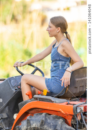 Young woman sits on minitractor and drives rural vehicle, performs loading work on farm 141064048