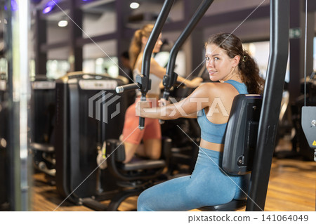 Positive young girl training on chest fly machine in gym Positive young girl training on chest fly machine in gym 141064049